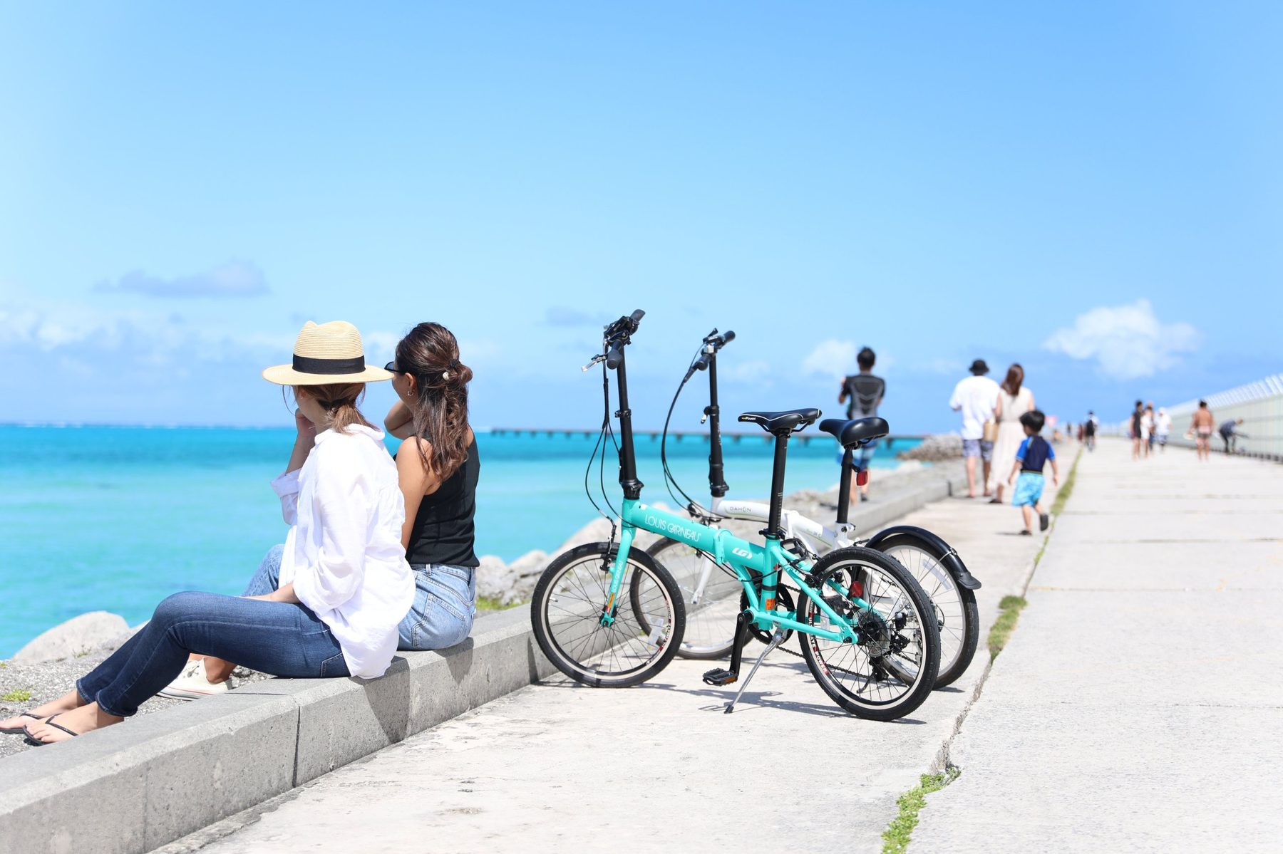 海沿いの防波堤に腰かけて海を眺める二人と、そばに停めたレンタサイクル